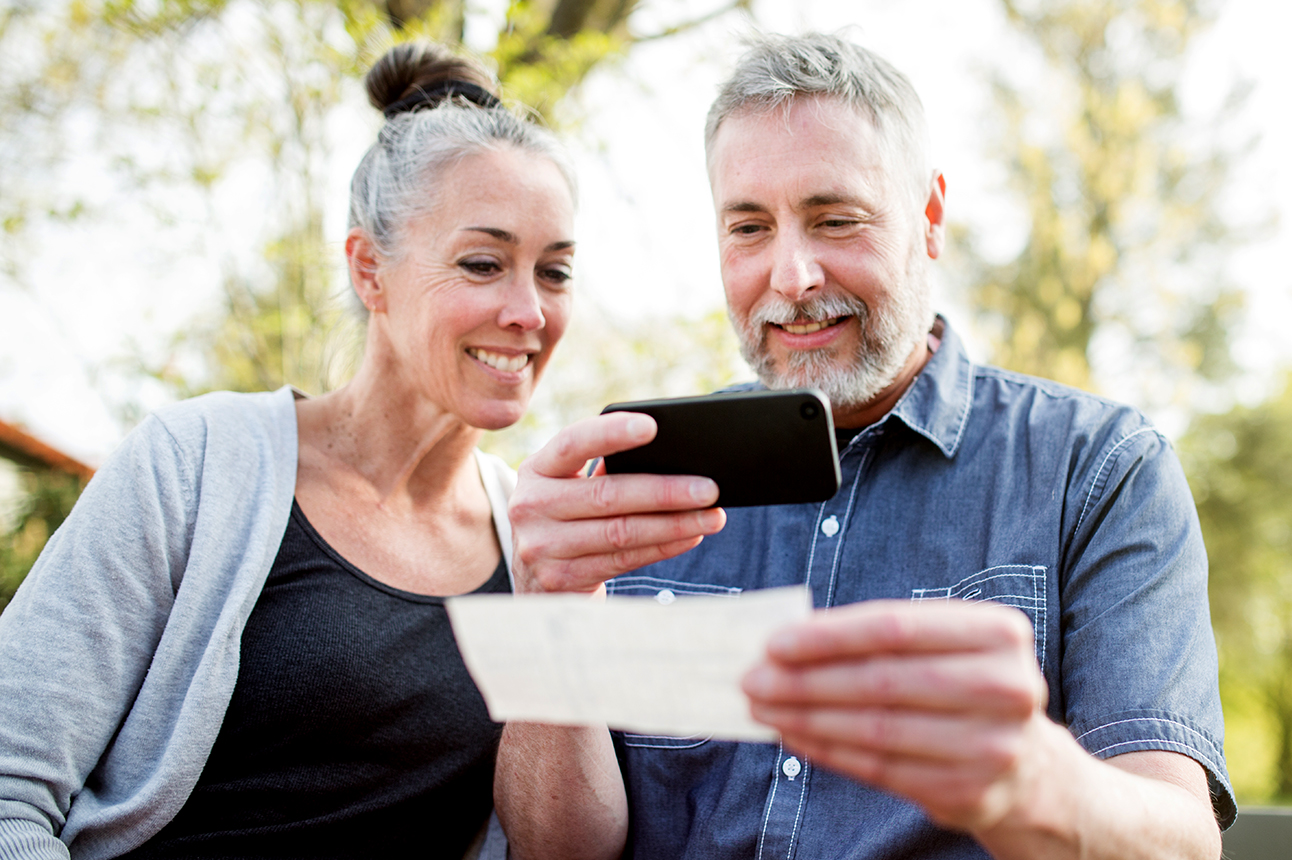 A gray haired and youthful looking couple in their 50's enjoy time with each other in a beautiful outdoor setting, the sun casting a golden glow on the scene. The man takes a picture of a check with his smart phone for a Remote Deposit Capture to his bank.